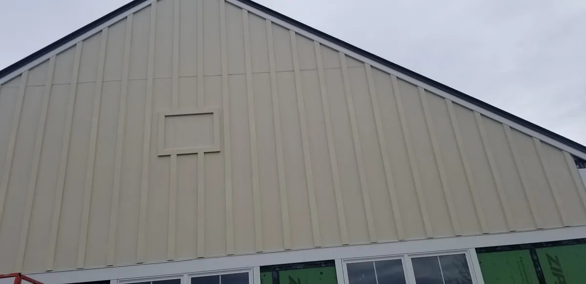 Tan board and batten siding covering a tall gable end on a new building, with windows and green sheathing visible along the bottom