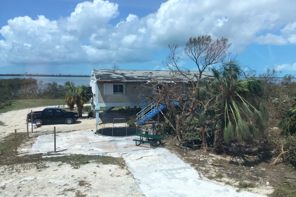Raised waterfront building with patches of shingles torn off its roof after a hurricane, with storm-stripped trees around it