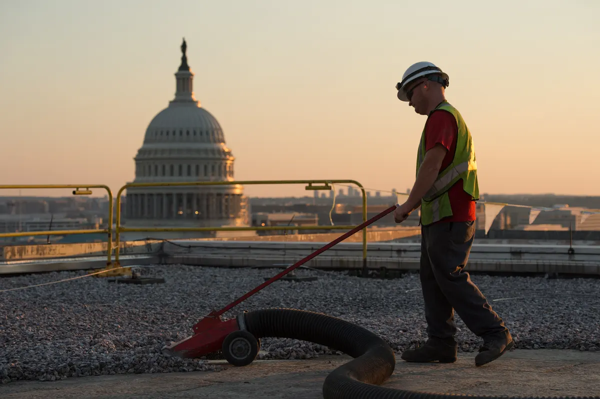 Roofer in a hard hat and safety vest vacuuming loose gravel ballast off a flat roof, with the US Capitol dome in the background at sunrise