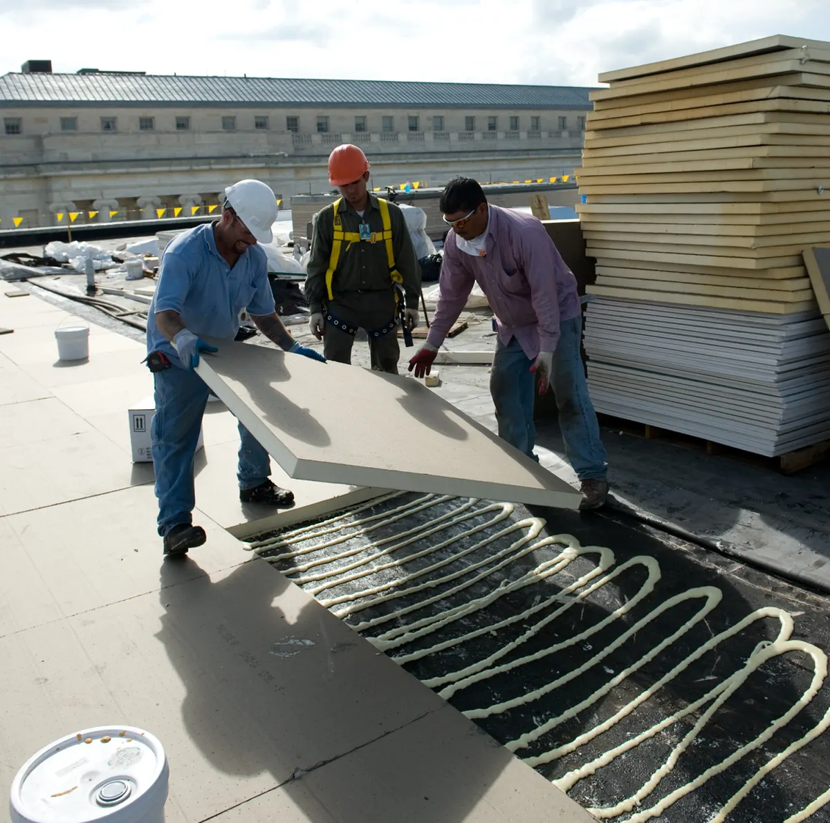 Three roofers in hard hats setting a new rigid insulation board into beads of adhesive on a flat roof, with stacked insulation boards beside them