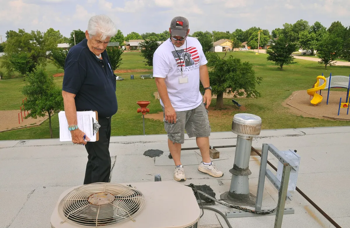 Two men inspecting a flat commercial roof beside a rooftop air conditioning unit, a vent pipe and patched membrane