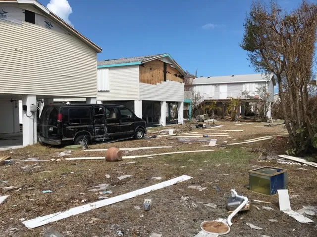 Raised homes after Hurricane Irma with vinyl siding torn off a gable wall, exposing the wood sheathing, and siding strips scattered across the yard