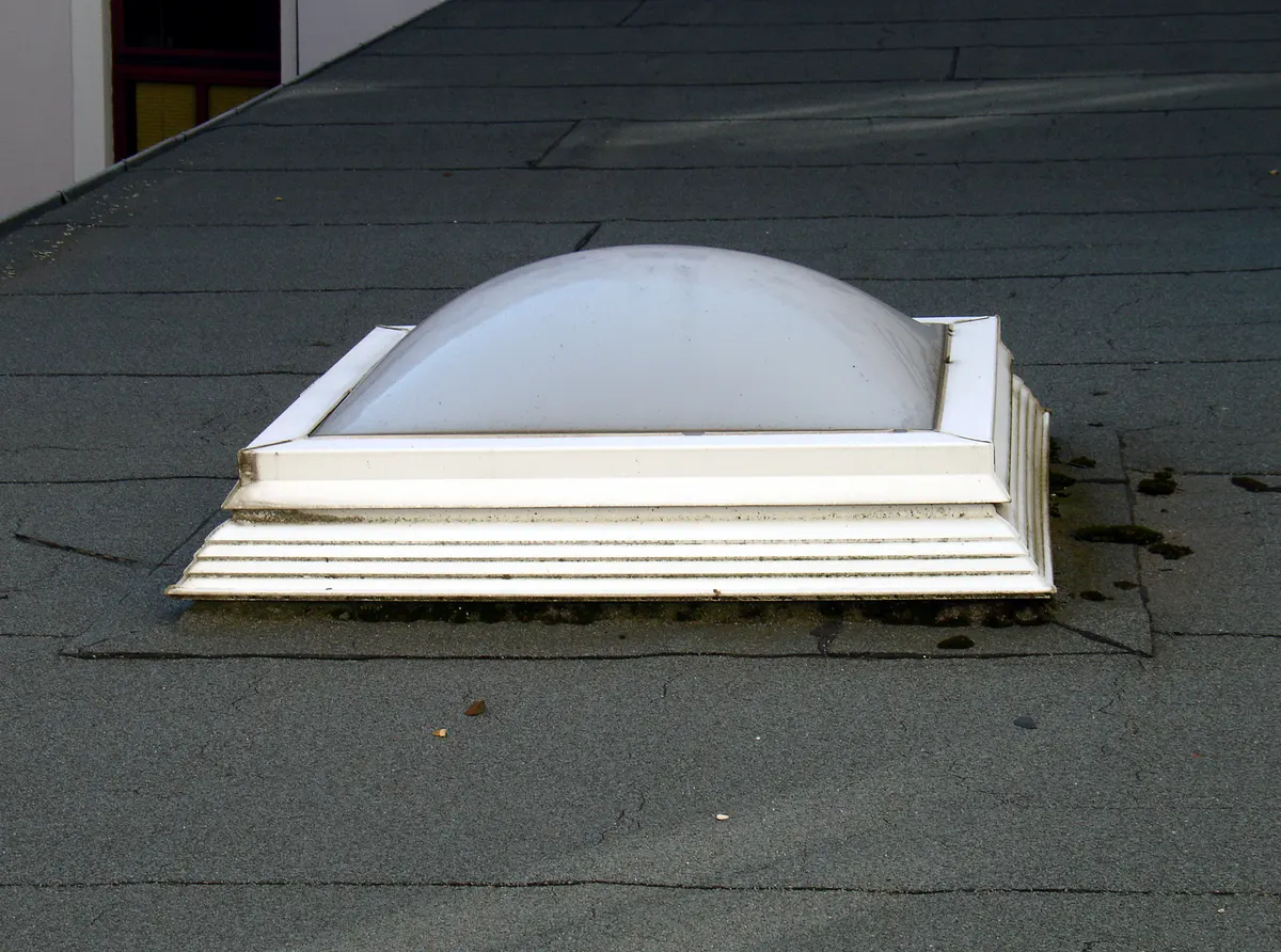 Older acrylic dome skylight on a raised white curb on a flat roof, with the dome clouded and the curb edges weathered